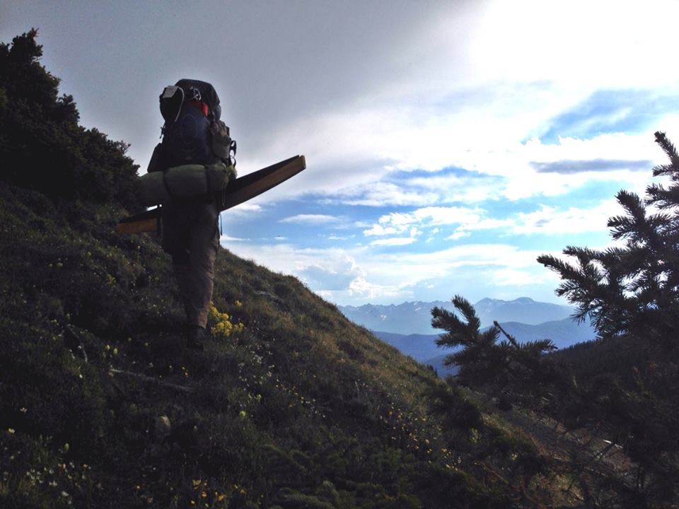 Hiker carrying a wooden board on a mountain trail, with distant mountains under a cloudy sky.
