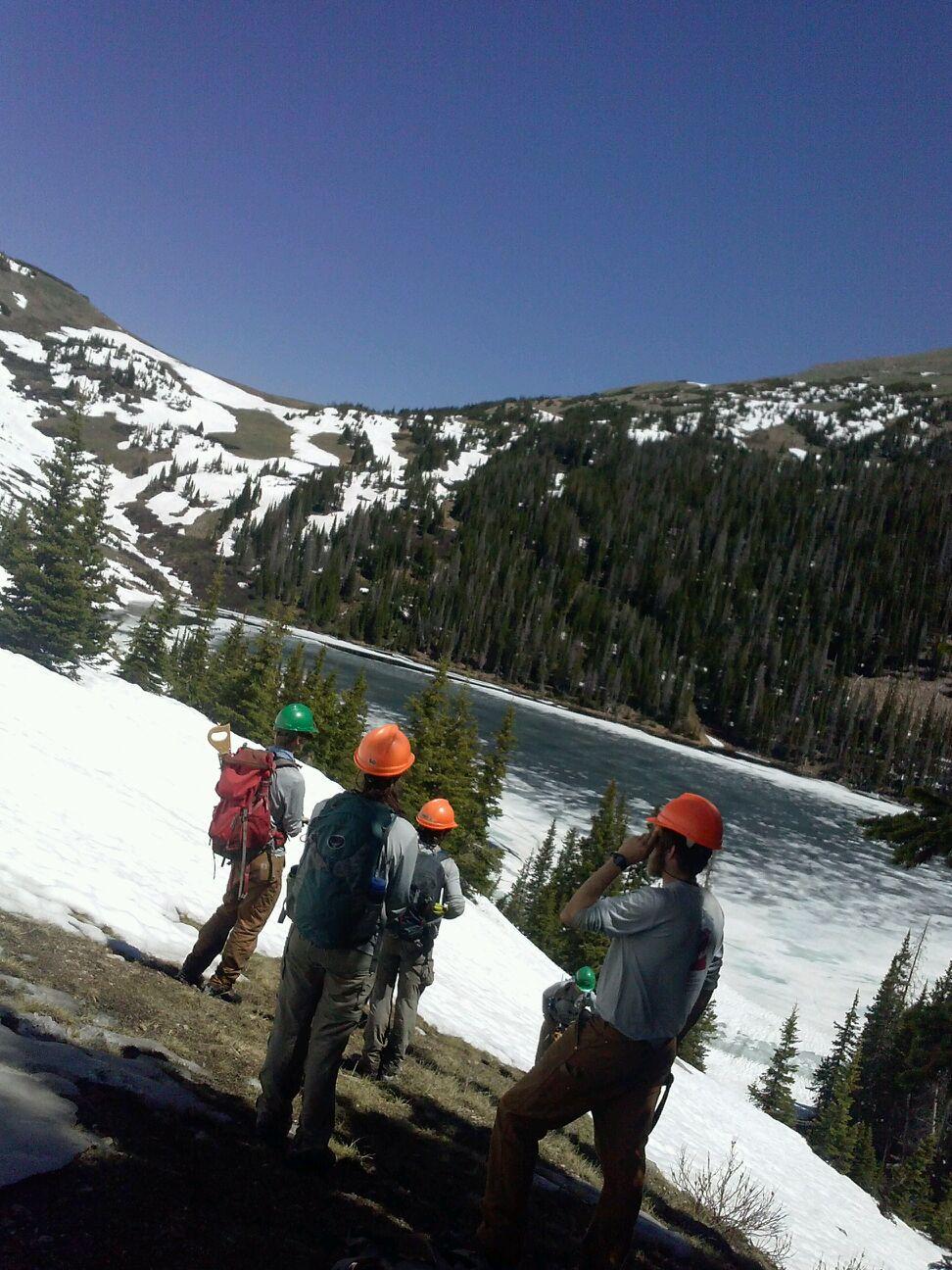 A group of hikers with backpacks standing near a snow-covered mountain lake