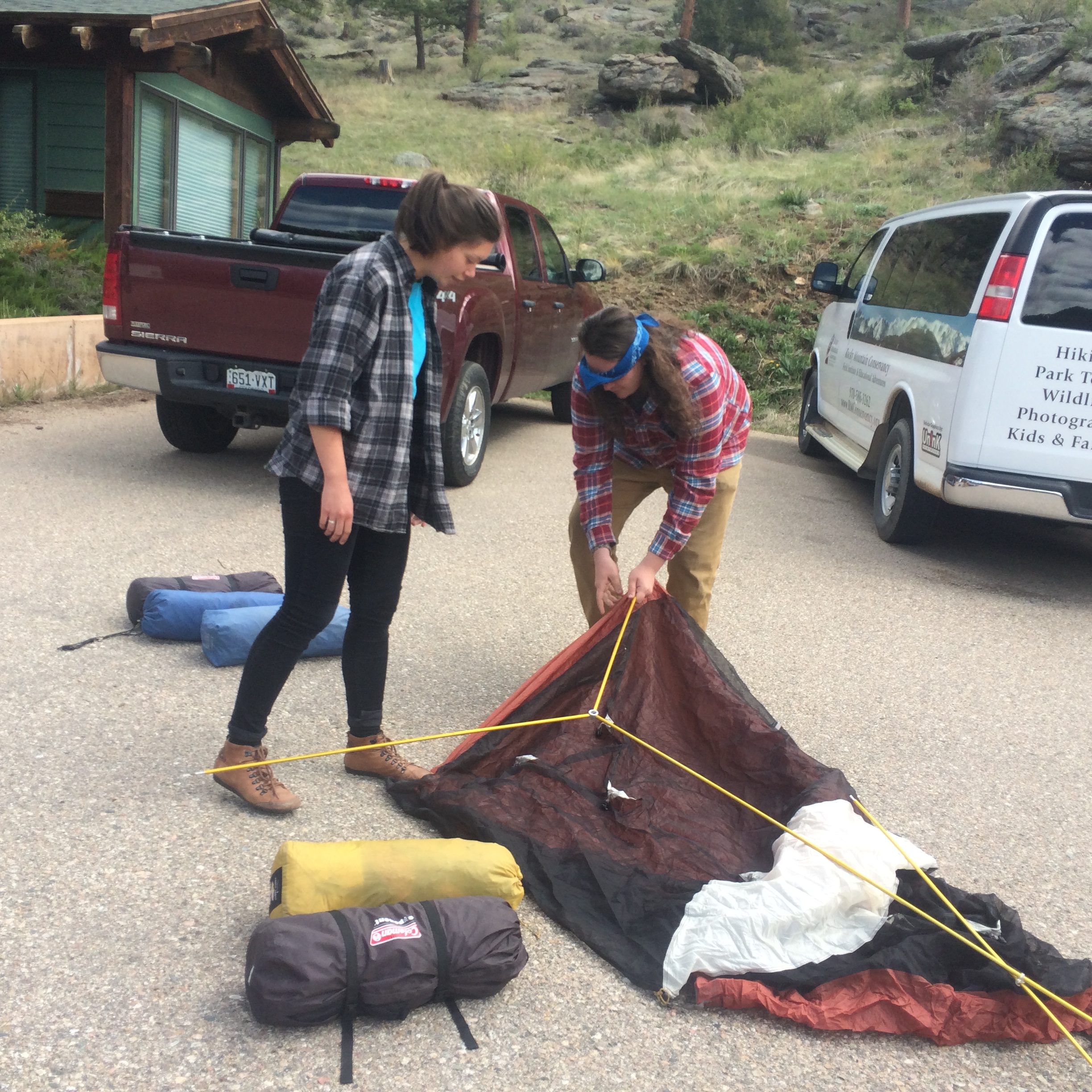 Two people setting up a tent near vehicles in a natural park area.