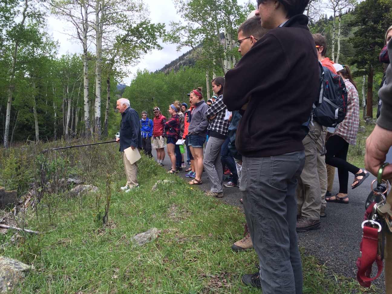 A group of people attentively listening to an elderly man holding a clipboard in a forested area.