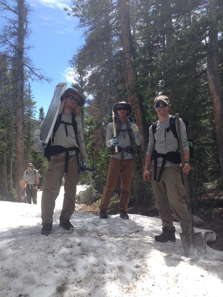 Three hikers with backpacks smiling on a snowy trail in a forest.
