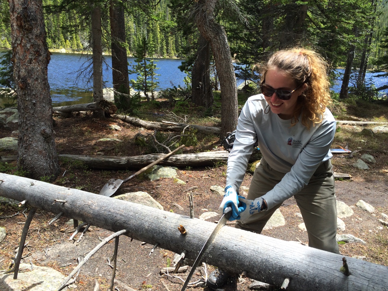 A woman wearing gloves saws a fallen log near a forest lake