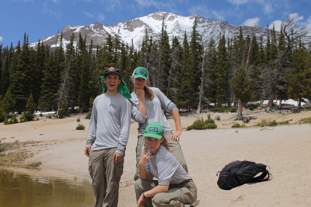 Three people stand by a lake with a snowy mountain in the background