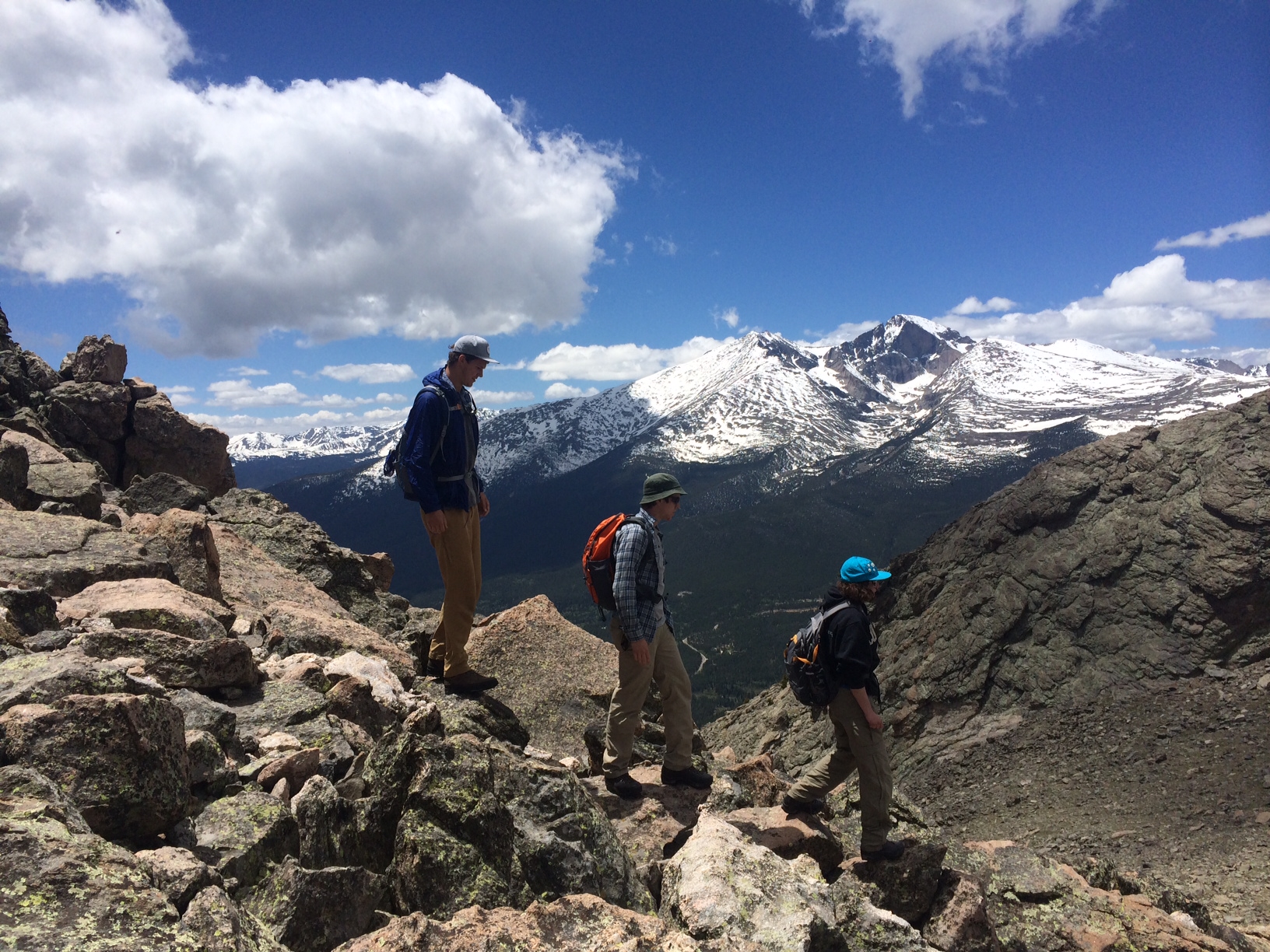Three hikers on a rocky mountain peak overlooking snow-capped mountains