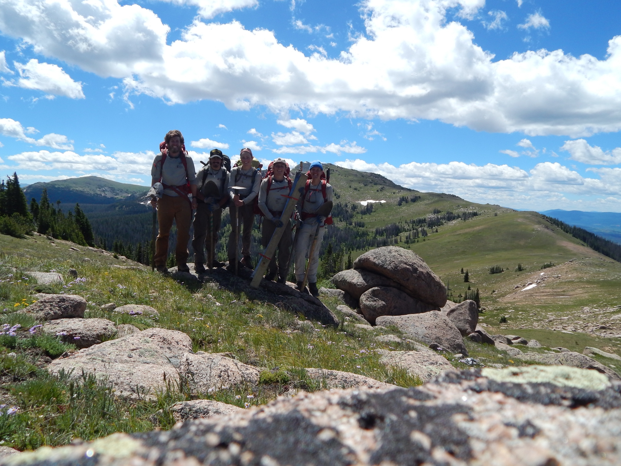 A group of hikers wearing backpacks posing together on a mountain trail with expansive views of hills and a partly cloudy sky.