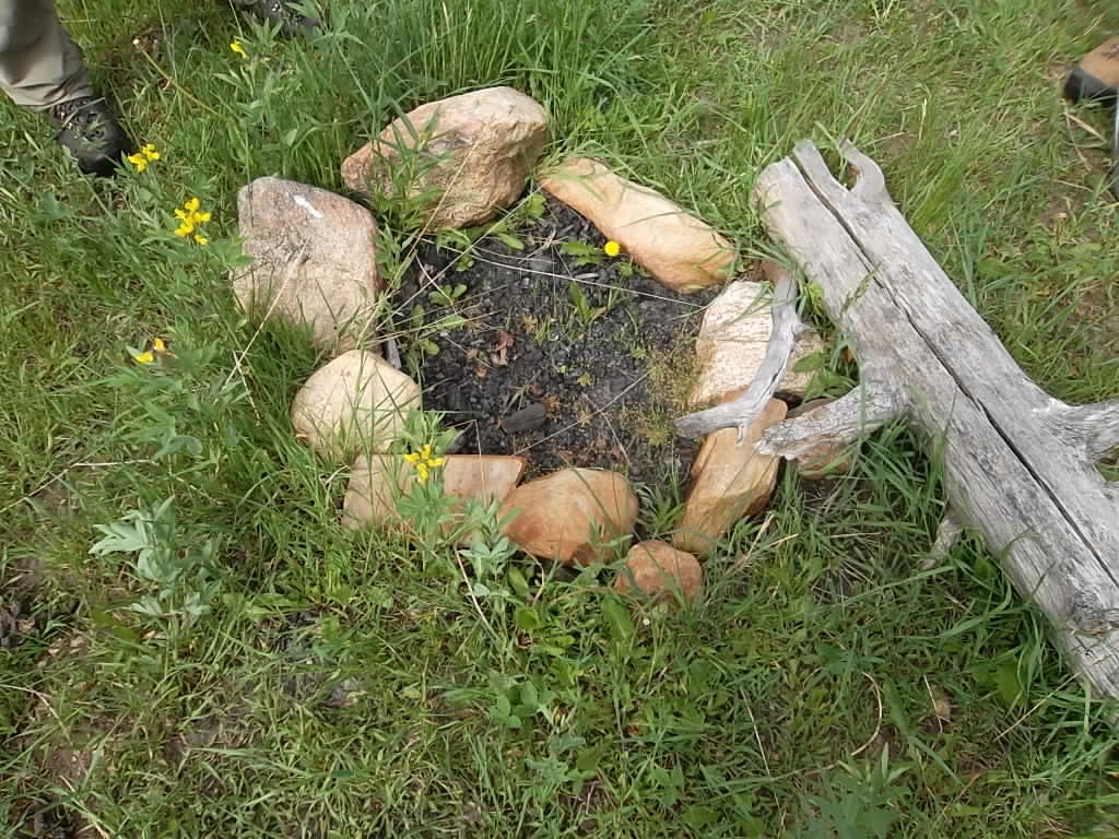 A simple, outdoor campfire ring made of stones, surrounded by grass and a few yellow flowers, with a person's boot visible on the edge.
