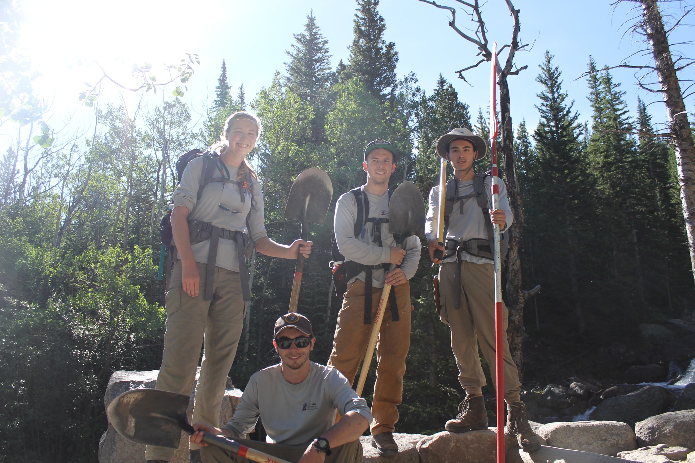 Four hikers with backpacks standing in a forest, smiling, with one kneeling in front. bright sunlight filters through trees.