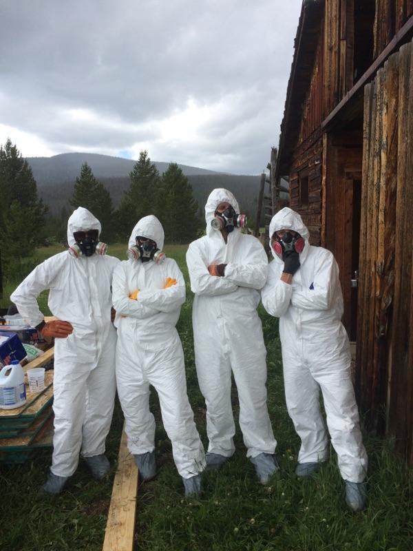 Four people in protective suits and masks standing beside a wooden building, with a forest and cloudy sky in the background.