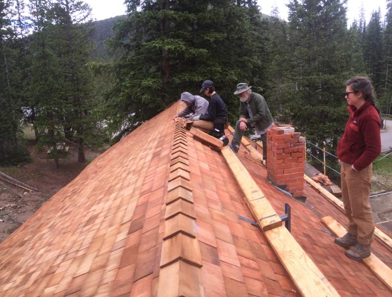 Three people work on laying shingles on a steep wooden roof, surrounded by forested landscape, while another person observes.