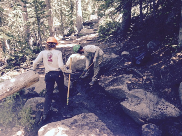 Two individuals wearing hard hats work on a rocky forest trail, one digging with a shovel and the other observing.