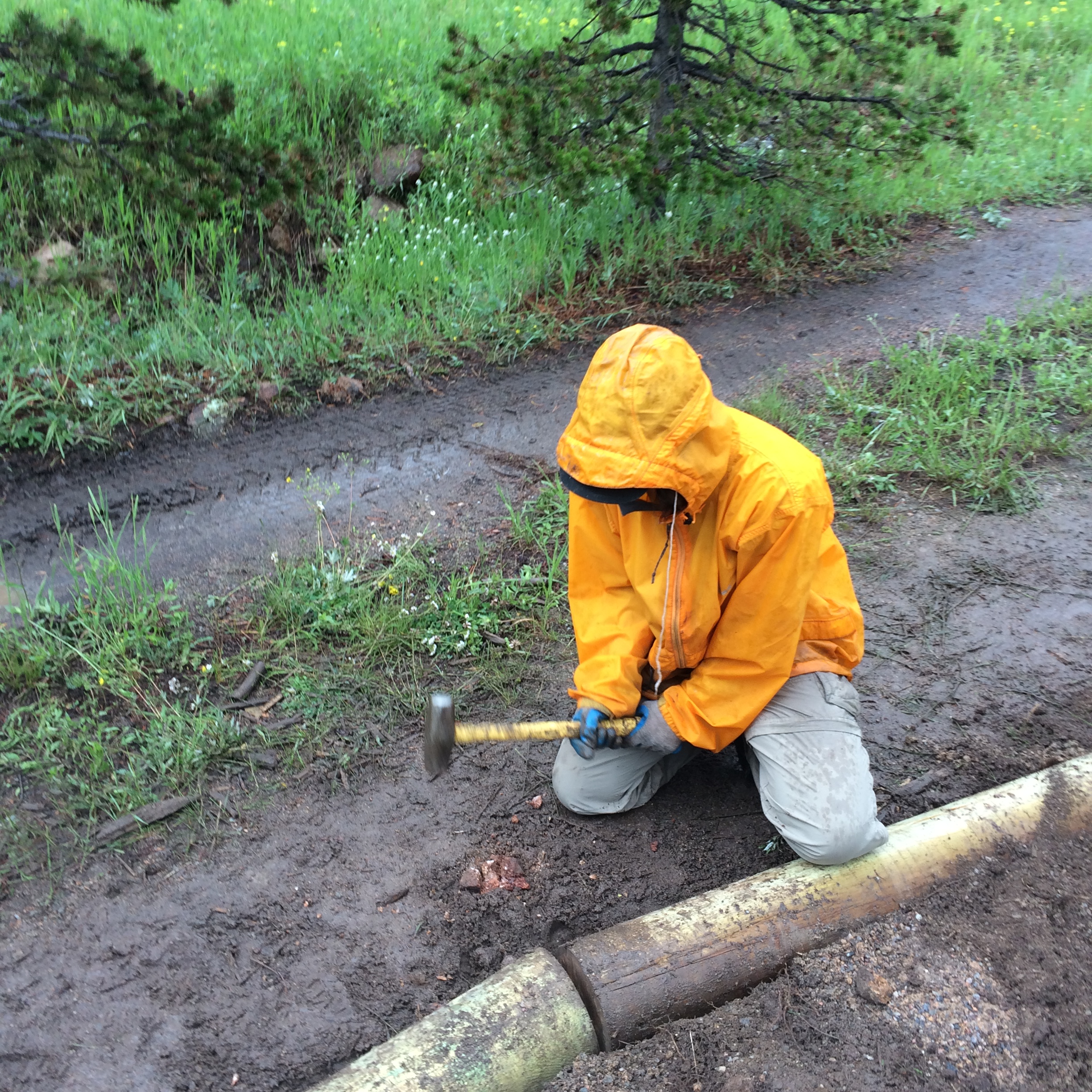 Person in orange raincoat using a hammer to secure a wooden log on the ground.