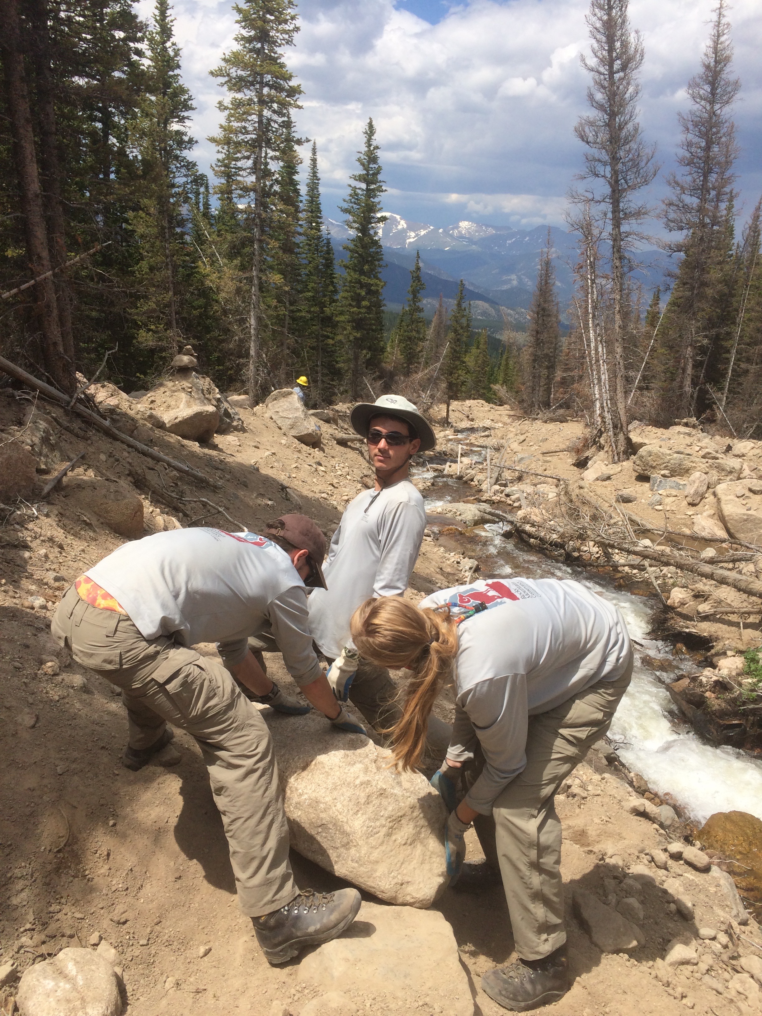 Three people in work uniforms lifting a large rock on a mountain trail, with pine trees and distant snow-capped mountains in the background.