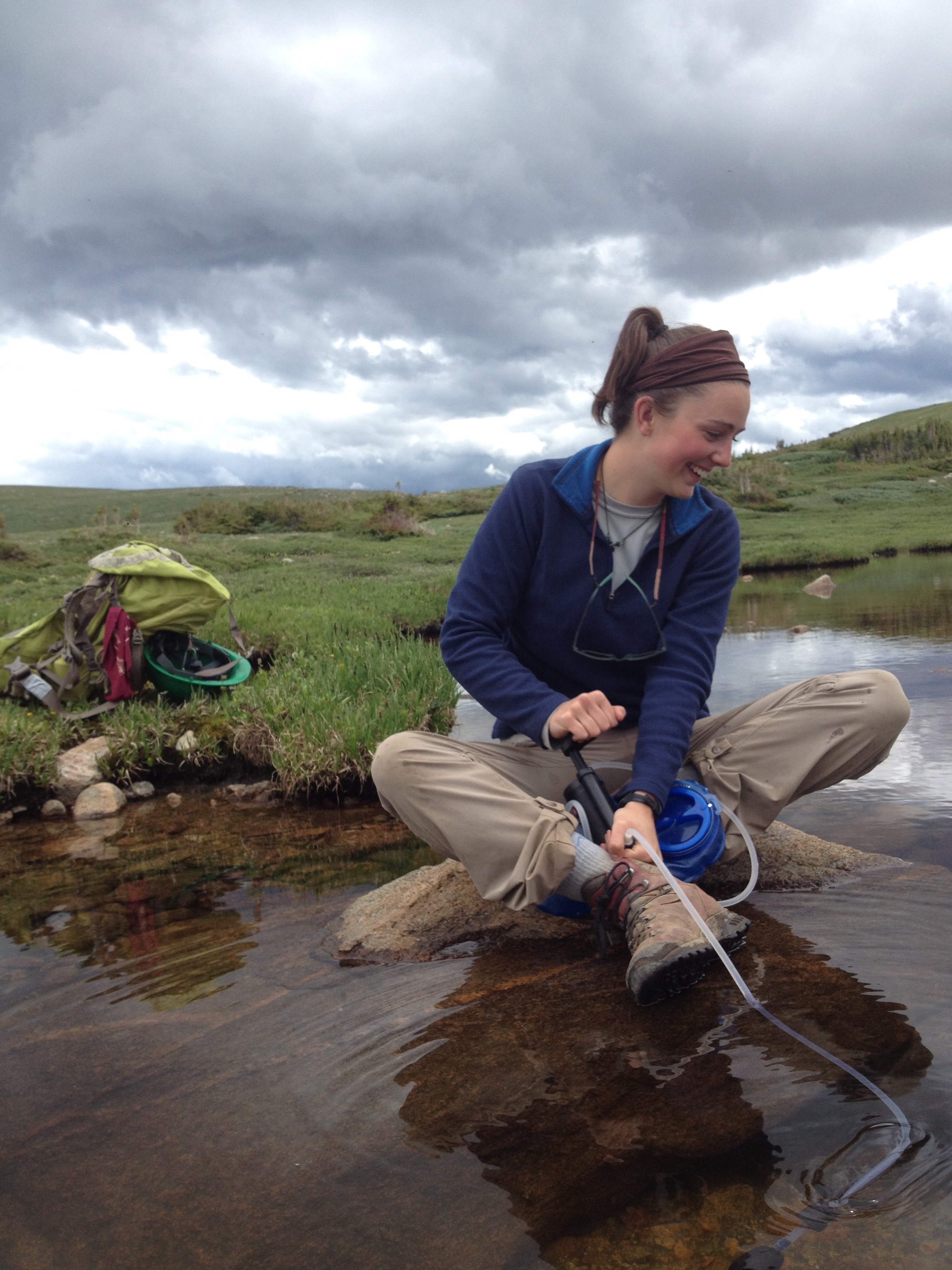 A woman using a portable water filter at a mountain stream, with a backpack nearby, under a cloudy sky.
