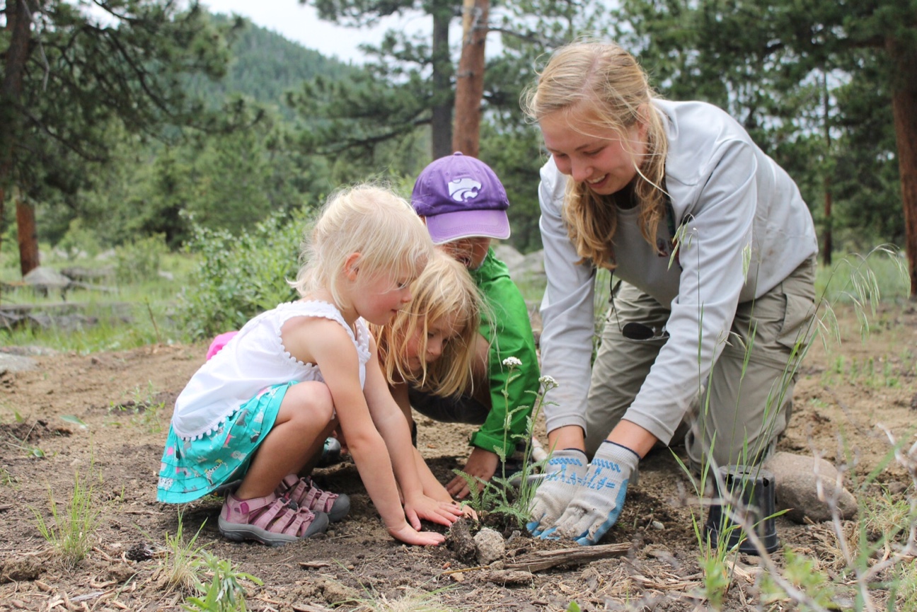 A woman and three young children examine soil and plants outdoors in a forested area, engaging in a learning activity.