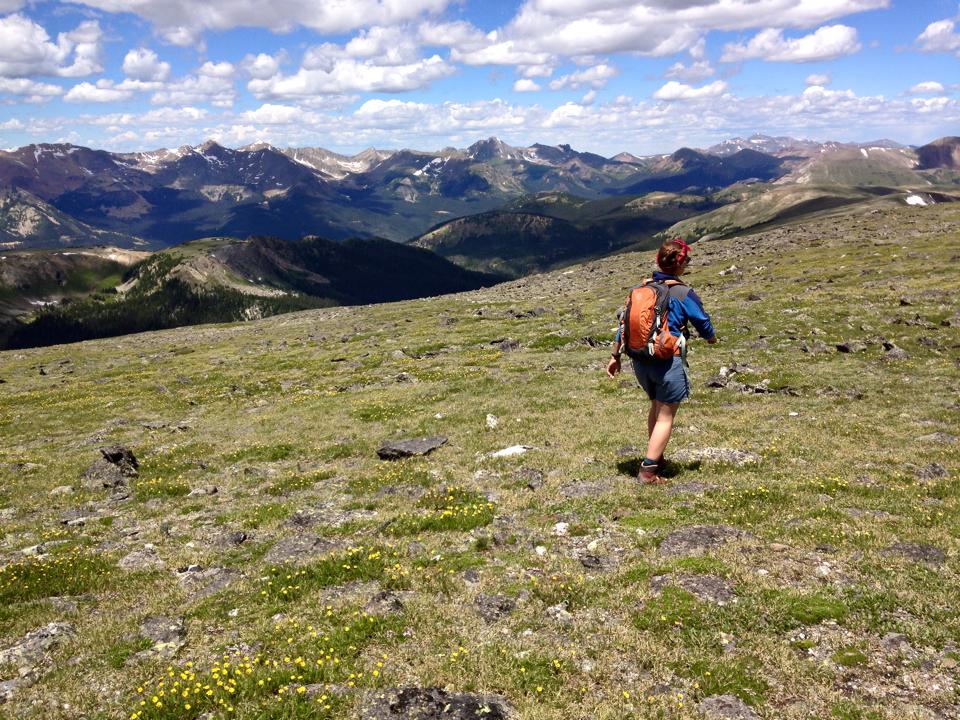 A hiker with a backpack standing on a grassy mountain slope, gazing at a vast range of distant mountains under a blue sky with clouds.