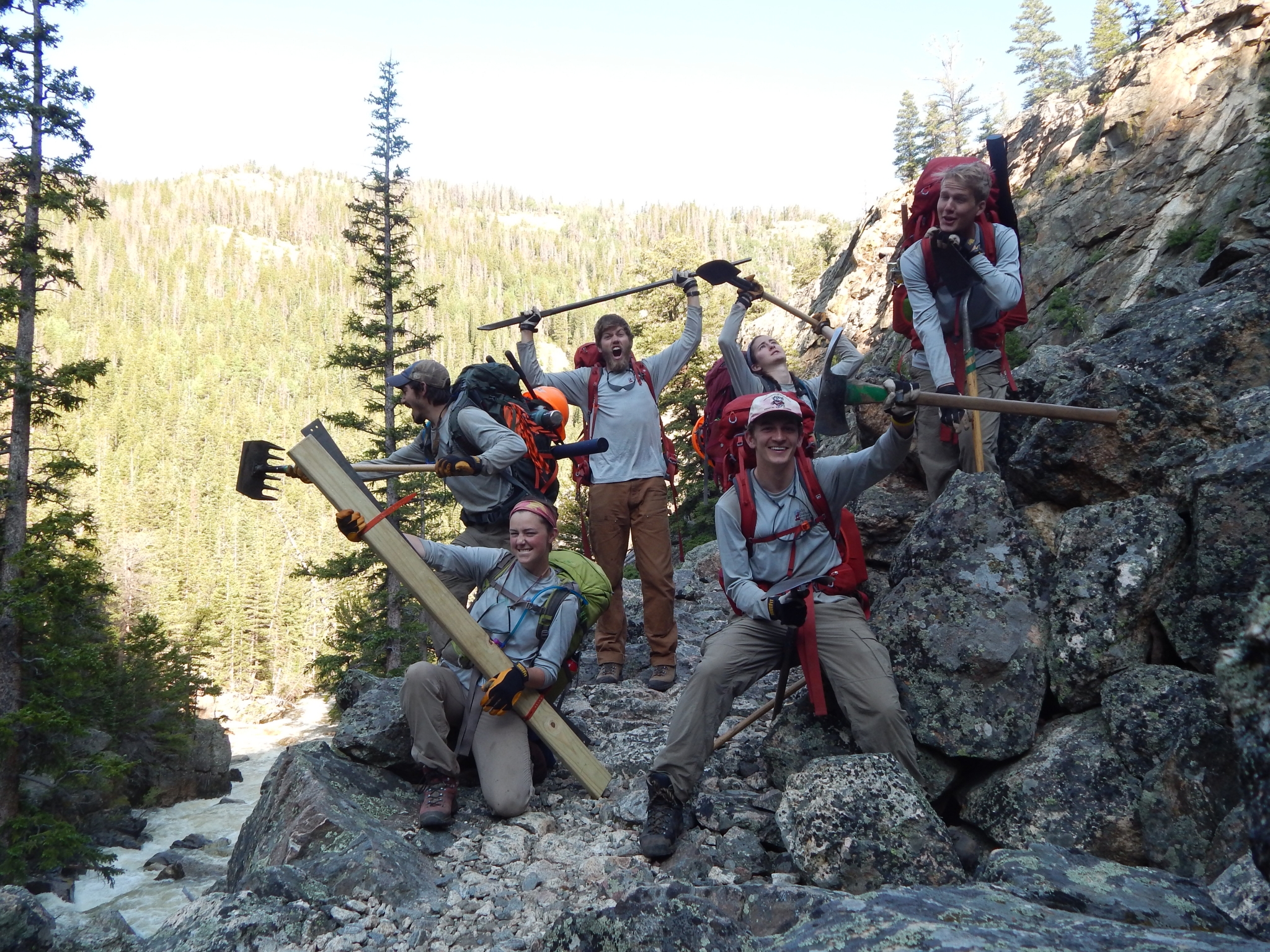 Seven hikers with tools posing on a rocky trail in a forested mountain area