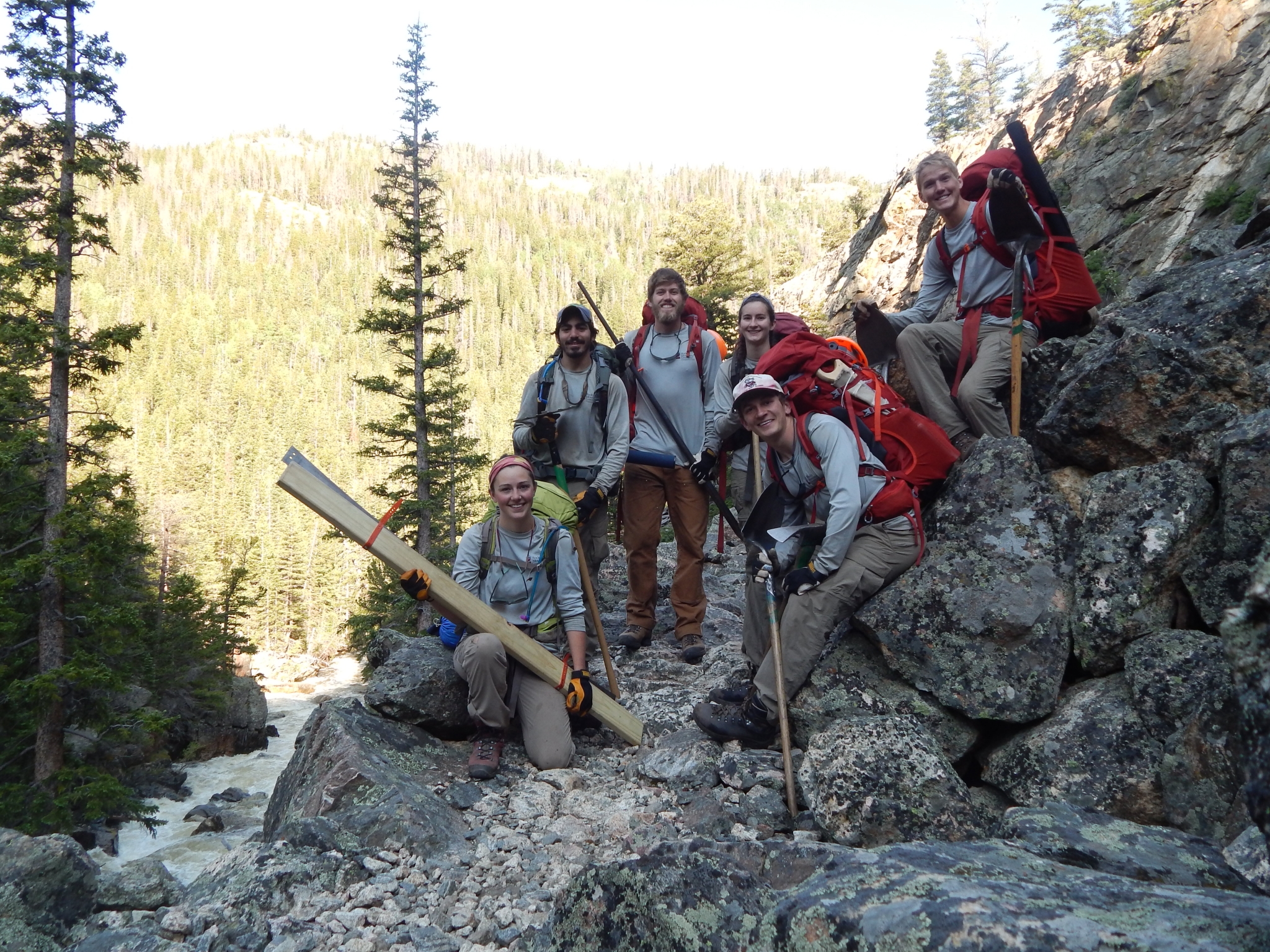 A group of seven hikers with backpacks posing on a rocky mountain trail surrounded by tall trees.