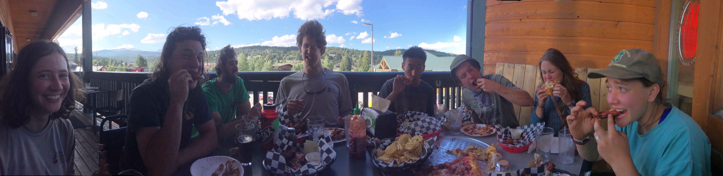 Group of friends enjoying a meal outdoors at a wooden table, surrounded by mountains under a blue sky.