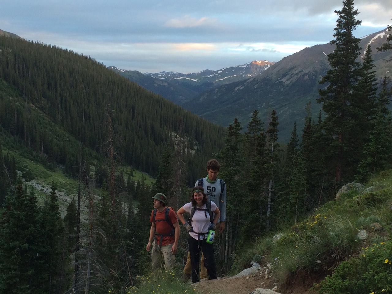 Three hikers on a mountain trail with lush greenery and distant snow-capped peaks during sunset.