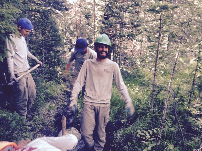 A man in a green helmet and safety goggles smiles at the camera while working outdoors with others