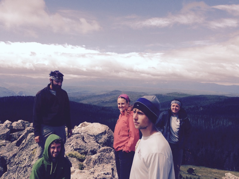 A group of five hikers at a mountain summit, with expansive forest views in the background under a cloudy sky.