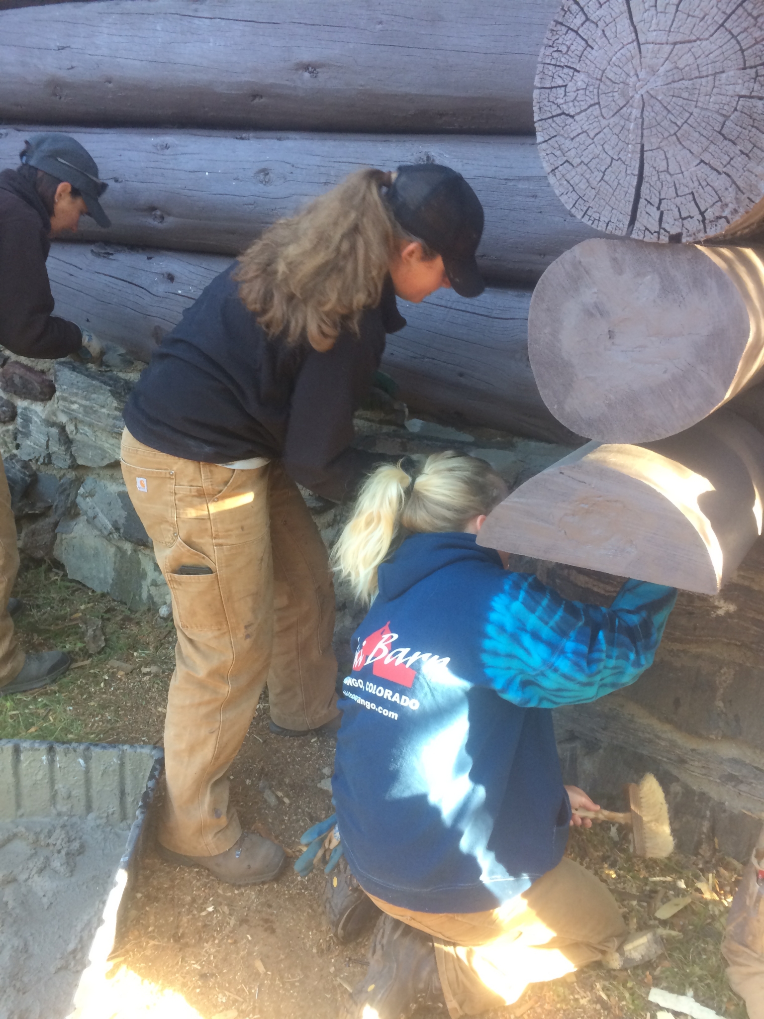 Three people, wearing work clothes and caps, inspecting a wooden structure outdoors in sunlight.