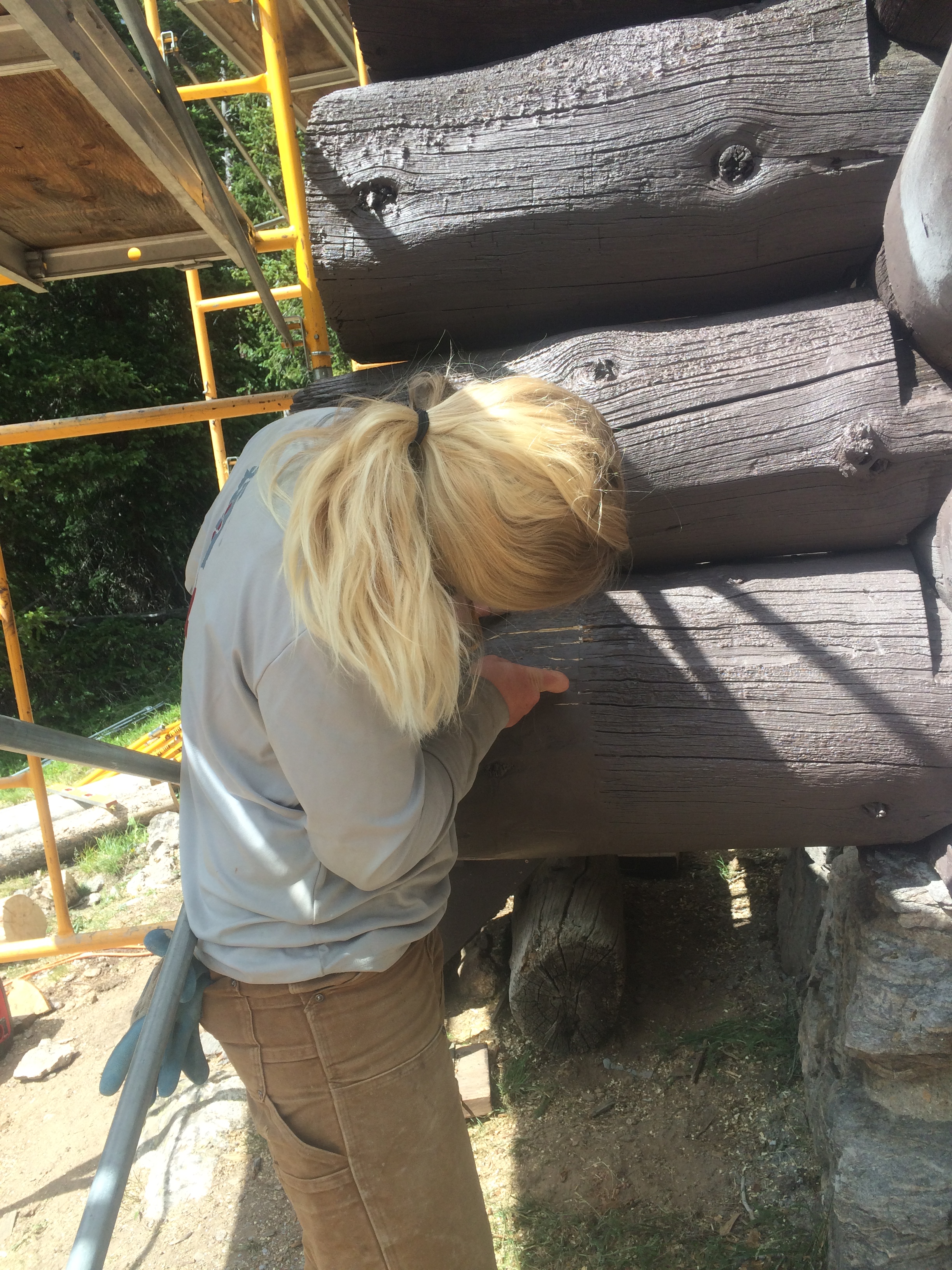 A person examining the wooden logs of a structure, with a yellow scaffold nearby.