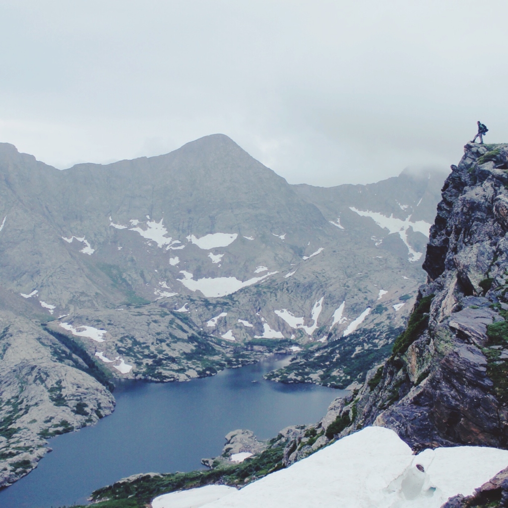A hiker stands on a rocky outcrop overlooking a mountain lake