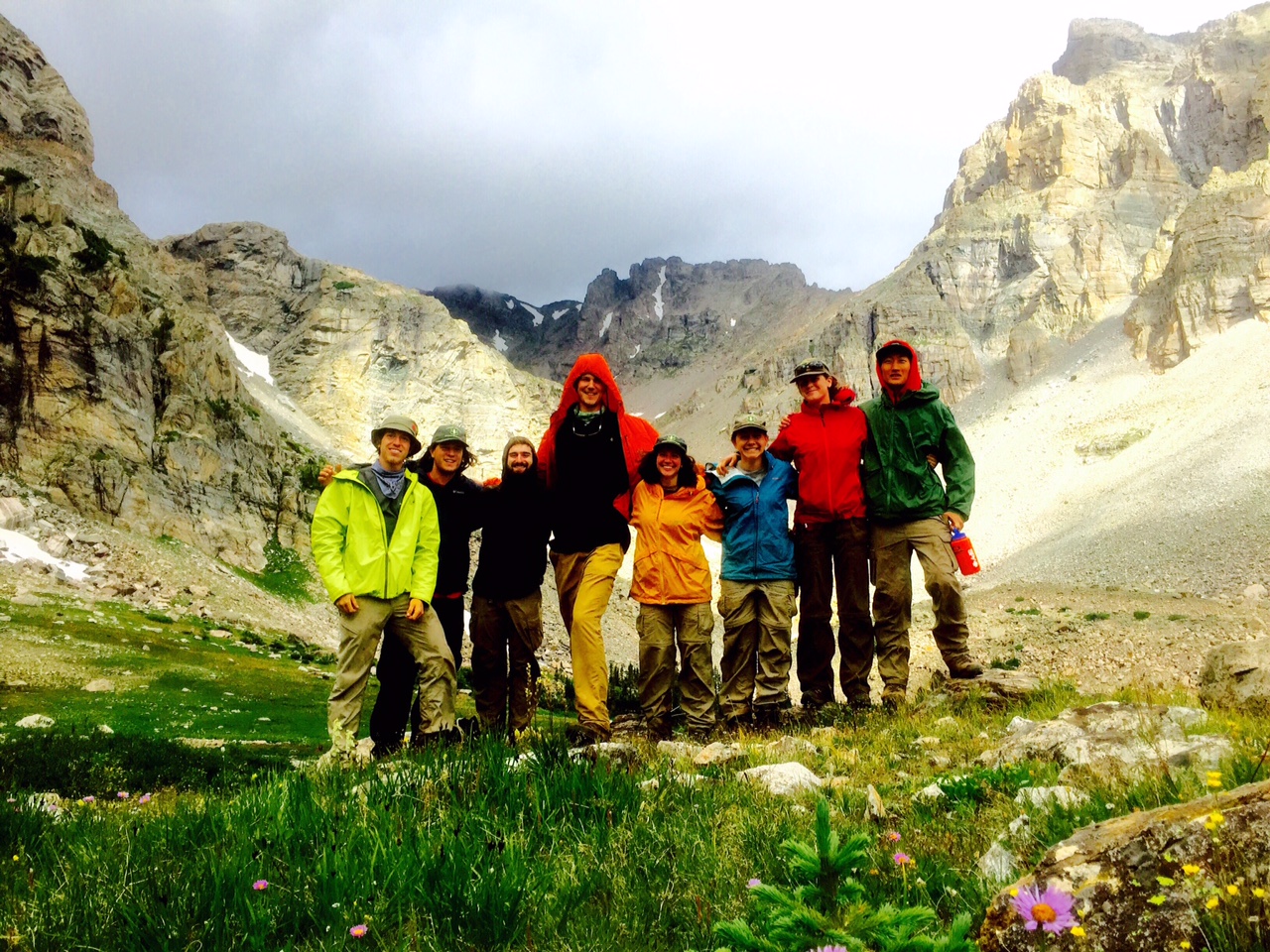 Eight hikers smile for a photo in a mountain meadow with towering cliffs in the background.