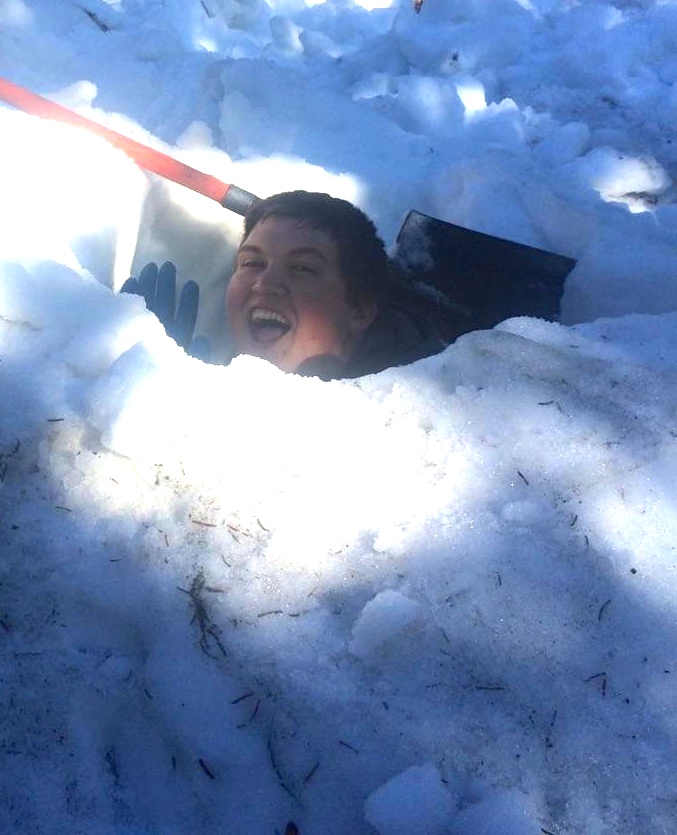 A person smiling while partially submerged in a snow hole, holding a red shovel.