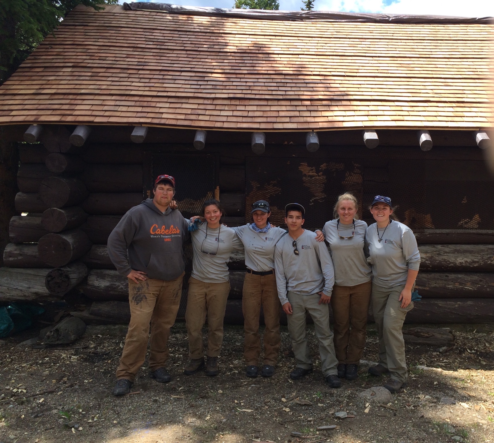Six park rangers in uniform standing proudly in front of a log cabin in a forested area.
