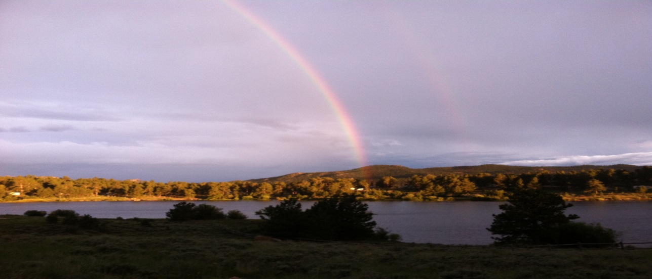 Double rainbow over a tranquil lake with forested hills and cloudy skies at dusk.