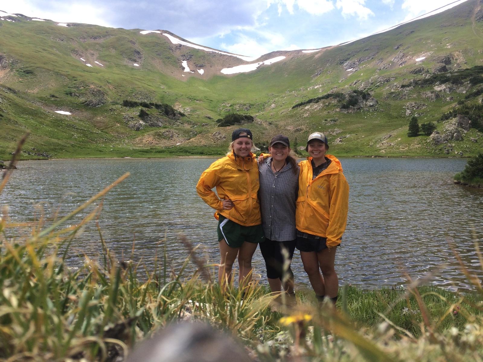 Three people in yellow jackets standing by a mountain lake with green hills and patches of snow in the background.