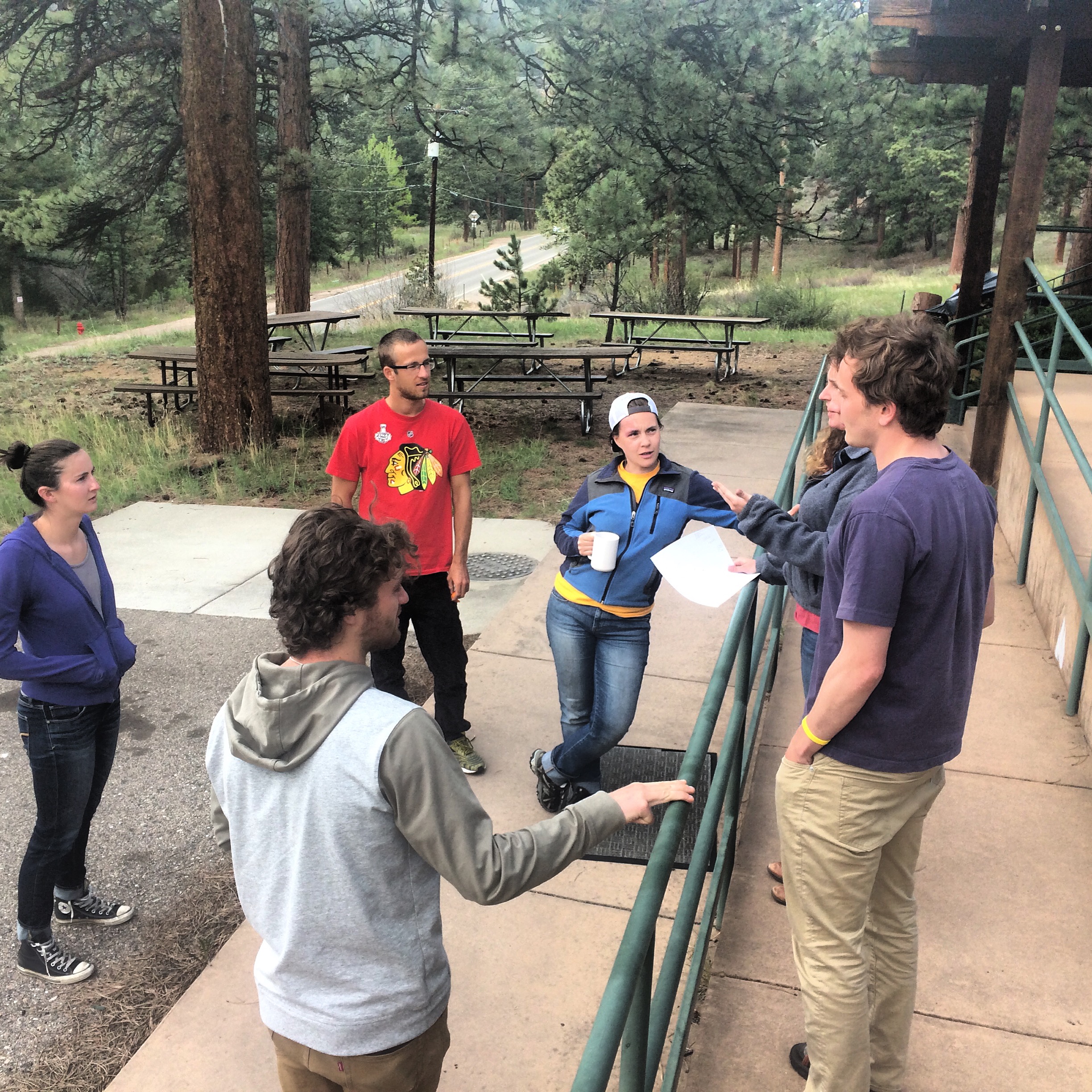 Group of six people engaging in a conversation outside a building, surrounded by trees in a park-like setting.