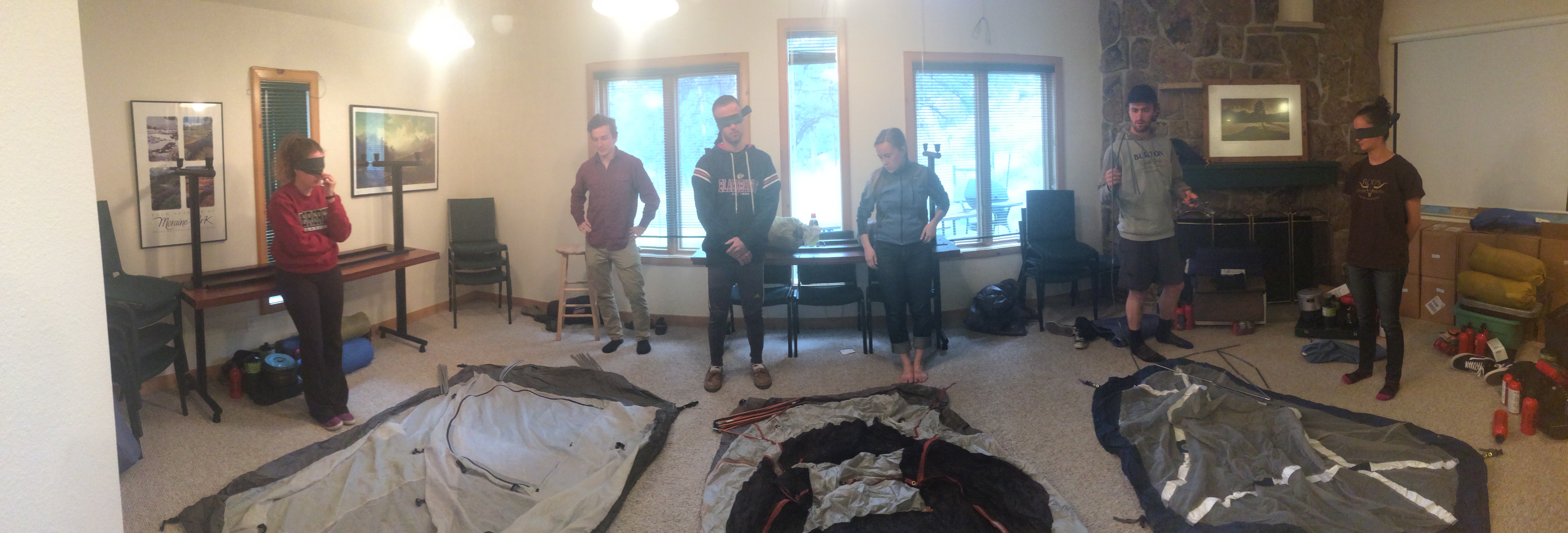Five people in a room with camping gear including a tent and backpacks, discussing their equipment amidst framed photographs on the walls.