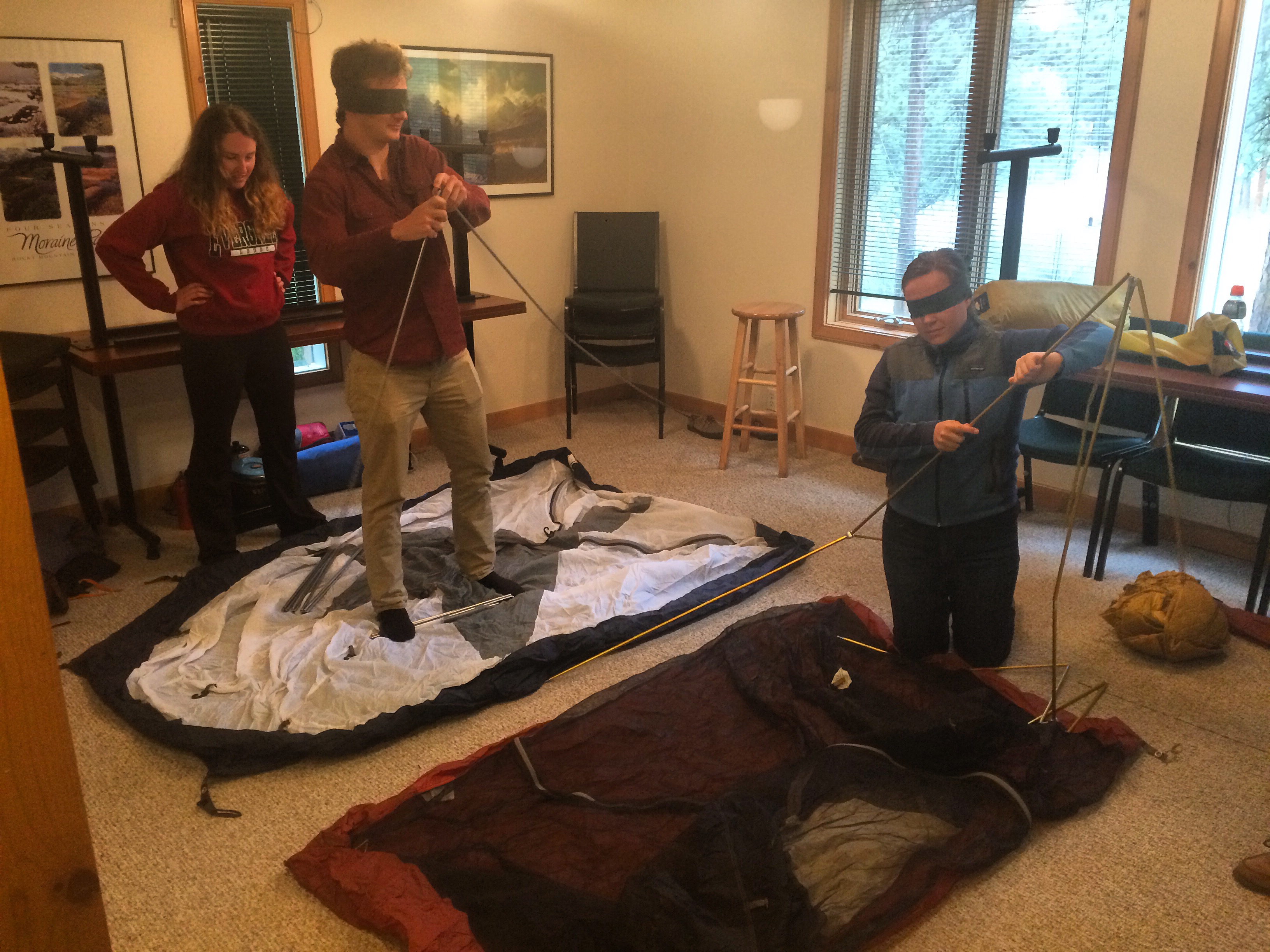 Three people assembling a tent indoors, with one man wearing a blindfold, another guiding him, and a woman observing.