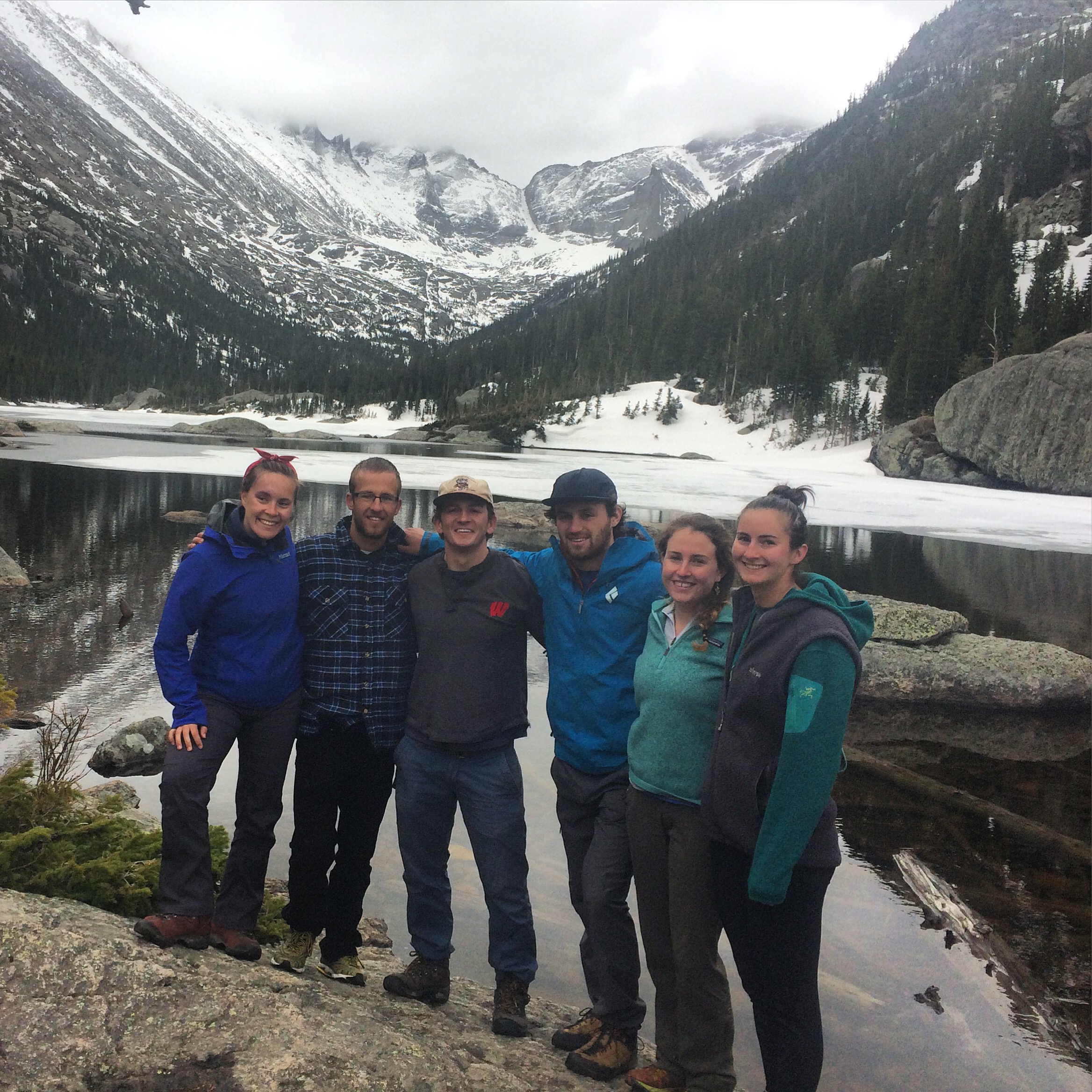 Group of six hikers smiling in front of a snowy mountain and partially frozen lake.