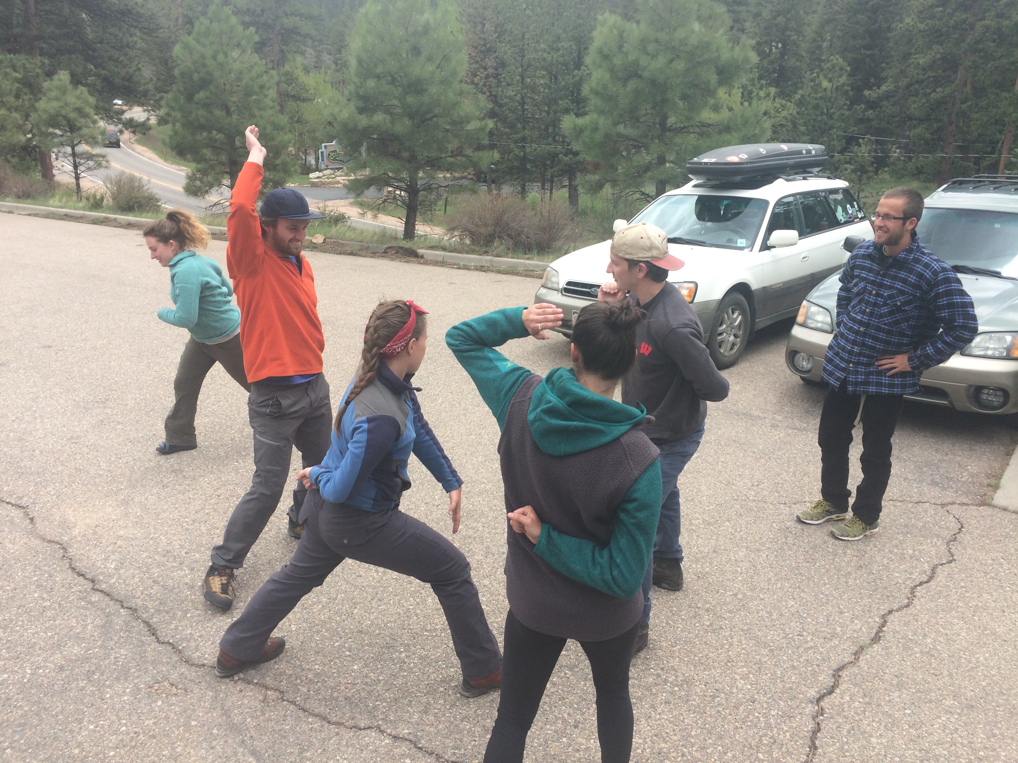 A group of adults playfully interacting in a parking lot, some in dance poses, with cars and trees in the background.