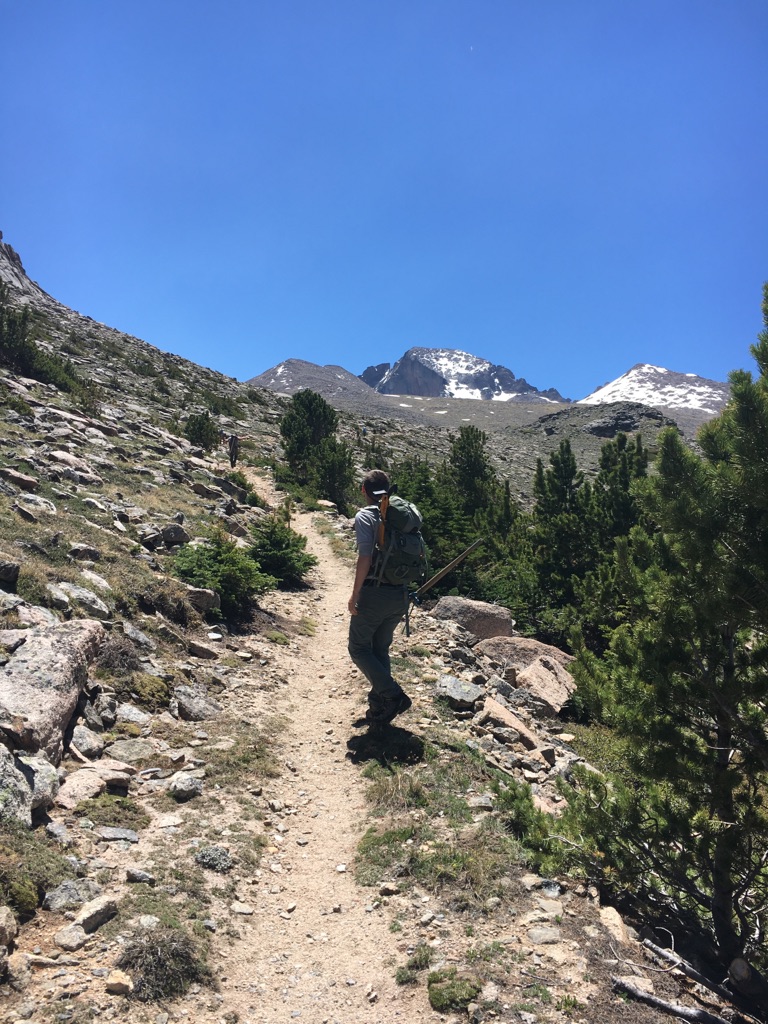 A hiker with a backpack and walking stick follows a trail towards distant mountains
