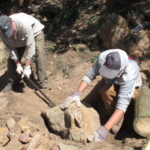 Two men digging a hole in the ground with a shovel.