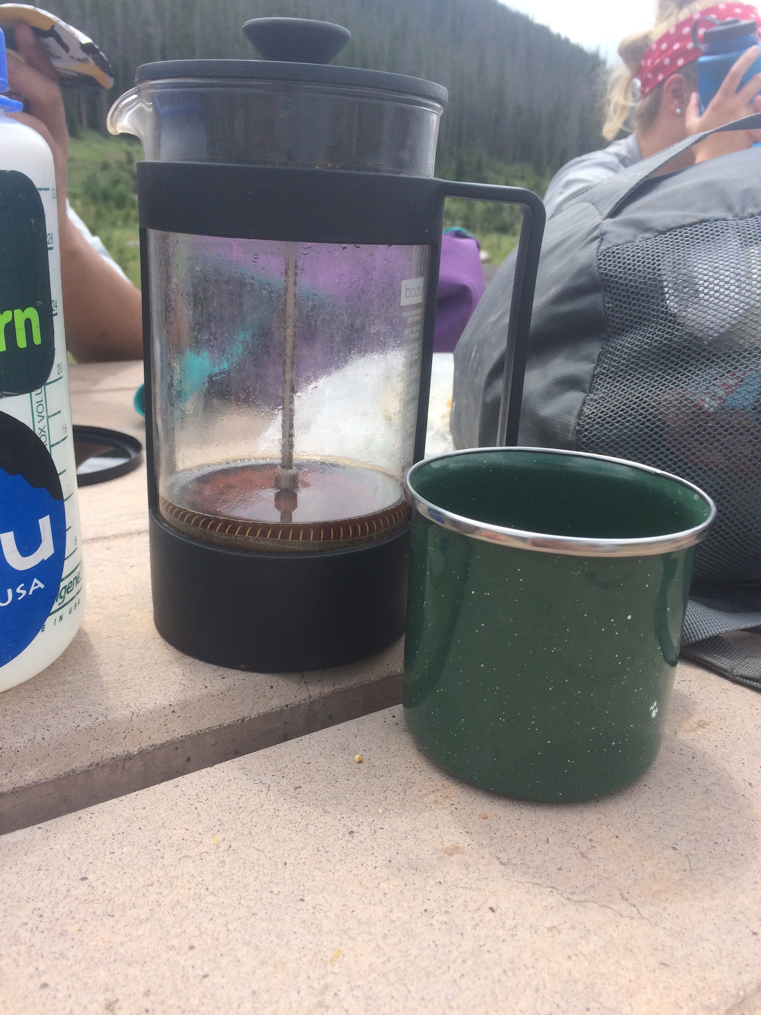 A french press coffee maker and a green camping mug on a wooden table outdoors