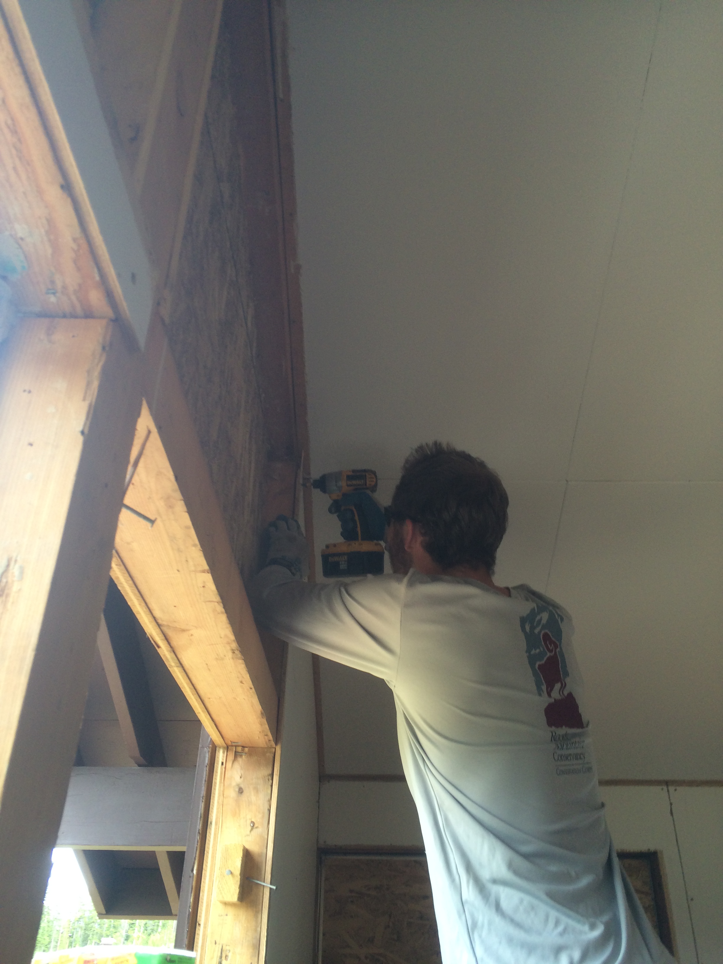 Man using a cordless drill to secure plywood to the ceiling joists in a construction setting.