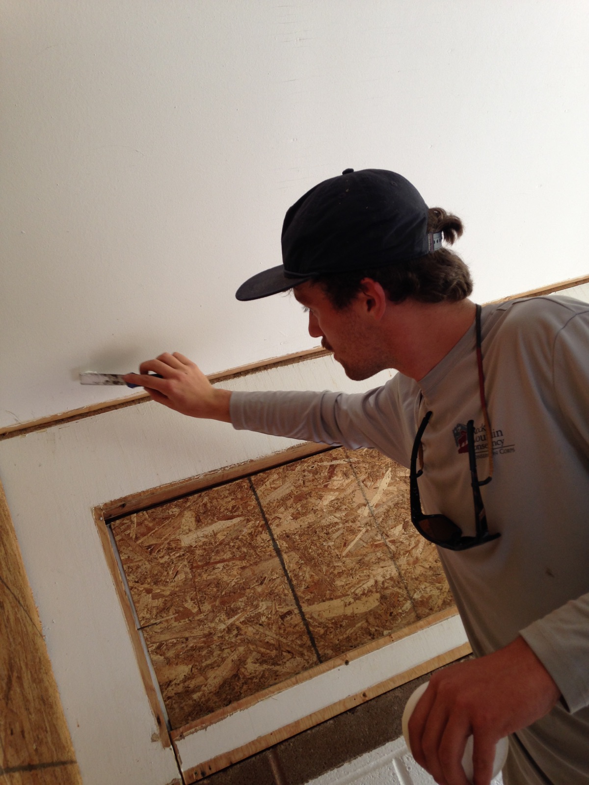 A man in a baseball cap uses a putty knife on a wall above a plywood panel.