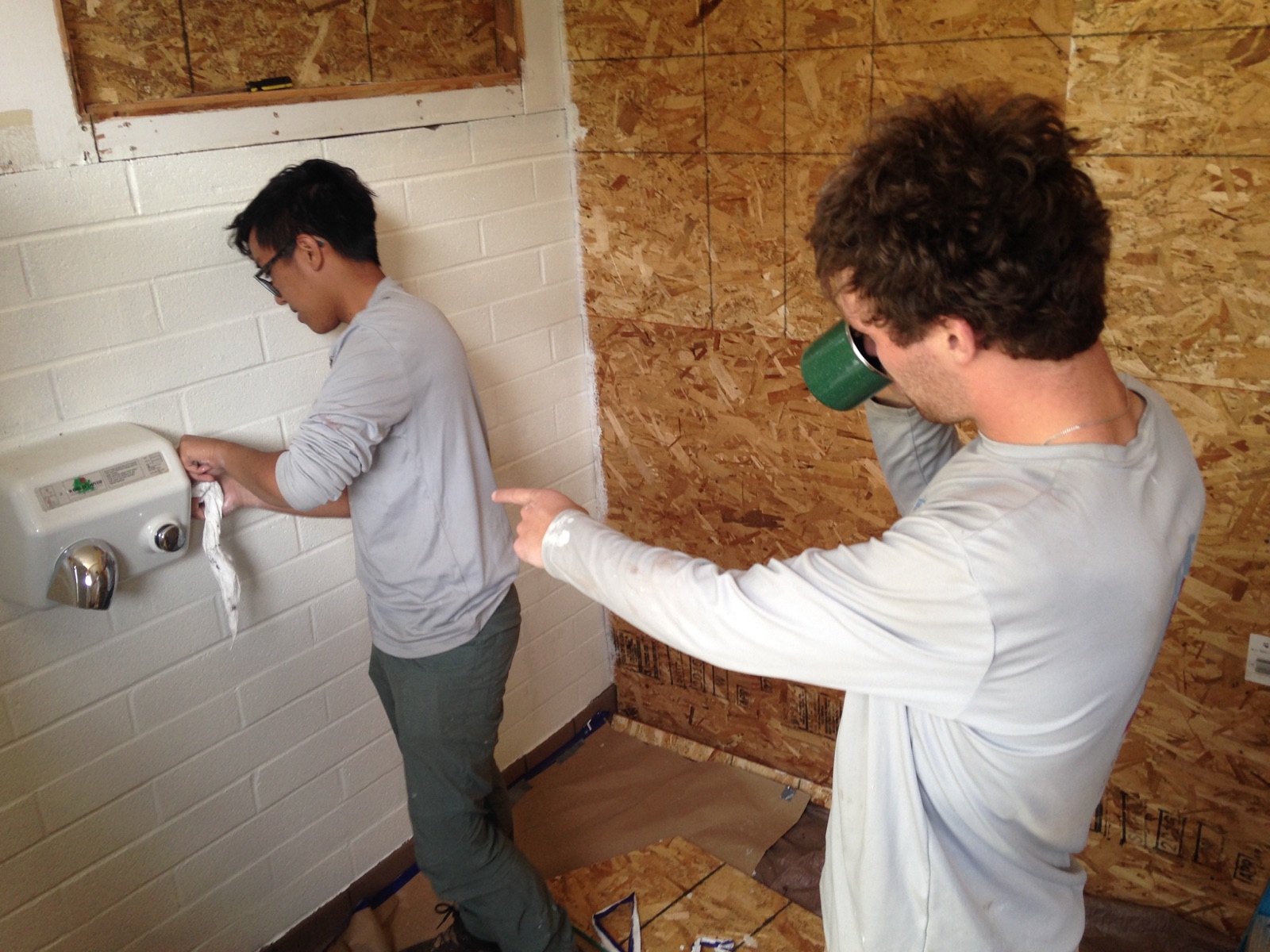 Two men working on a wall, one operating a hand dryer and the other drinking from a mug