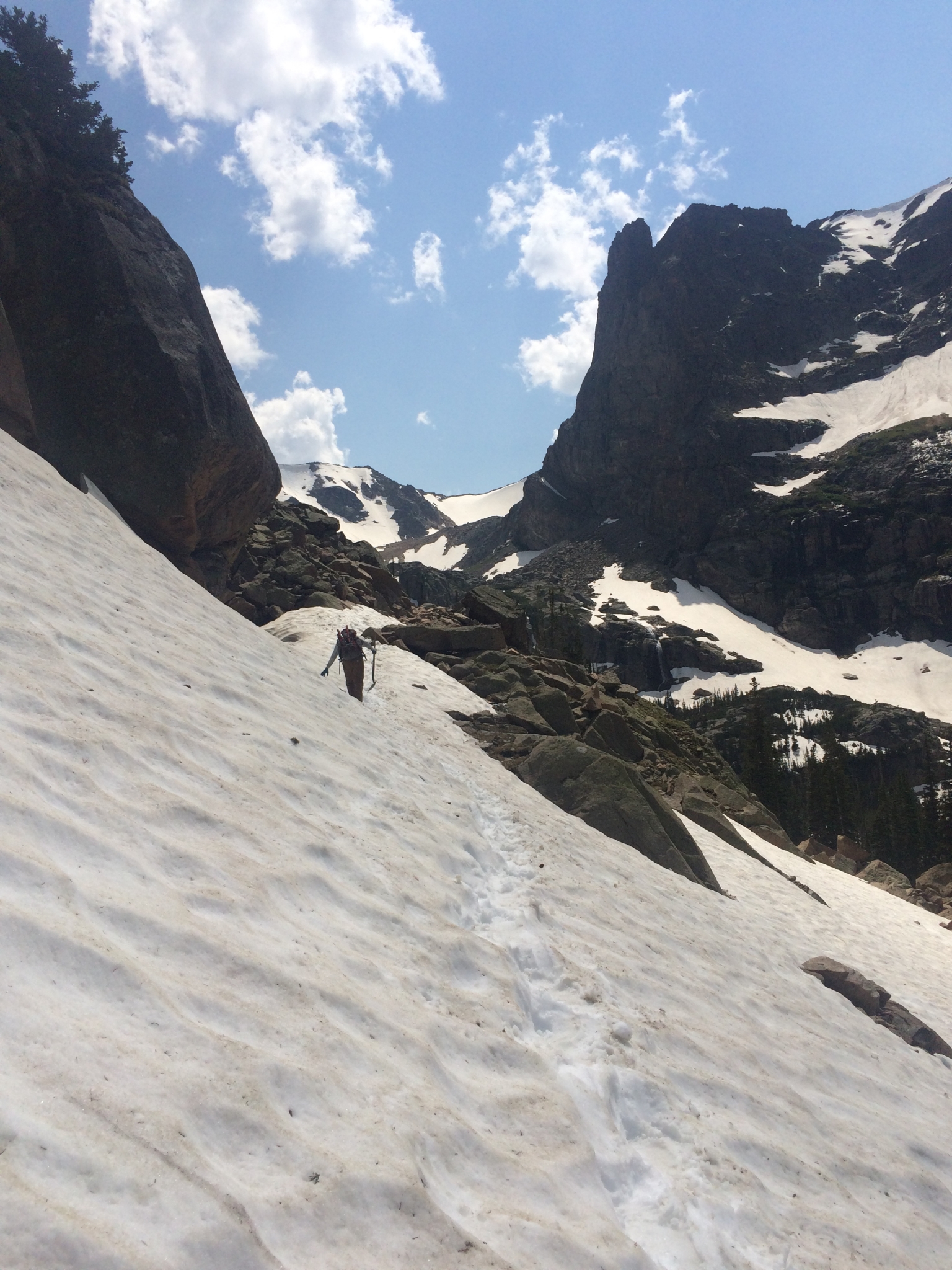A lone hiker ascending a steep snow-covered slope in a rocky mountainous area on a sunny day.