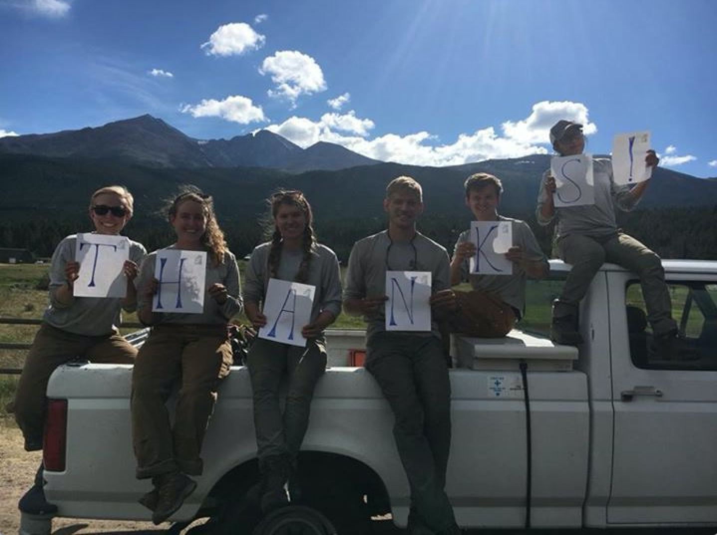 A group of seven people on a white truck display letters spelling "thanks"