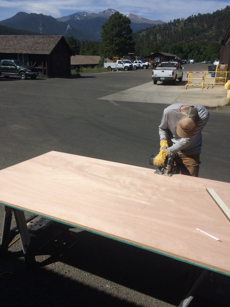 A worker cuts a large sheet of plywood with a circular saw in a parking lot