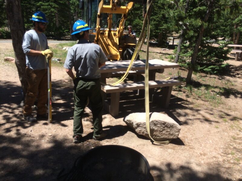 Two workers in hard hats using a crane to install a picnic table in a forested area.