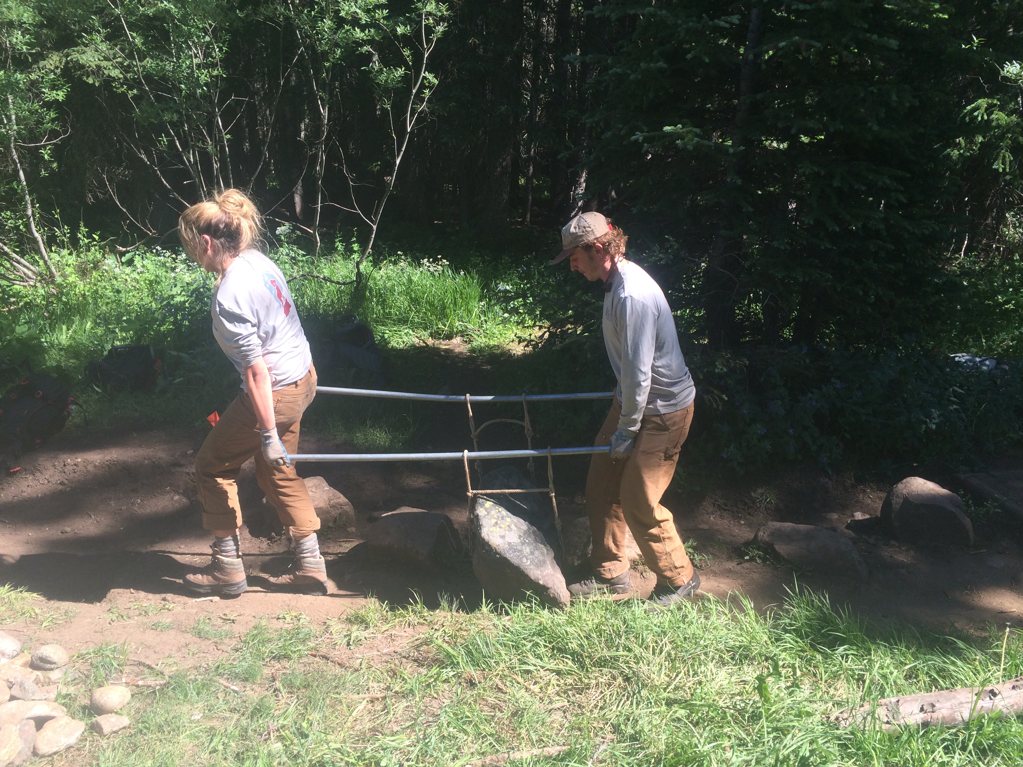 Two people using a long metal bar to lift a large rock in a forested area.