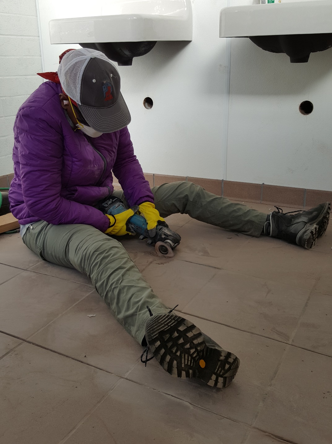 A person in a purple jacket sitting on a tiled floor, using a power tool unto the floor